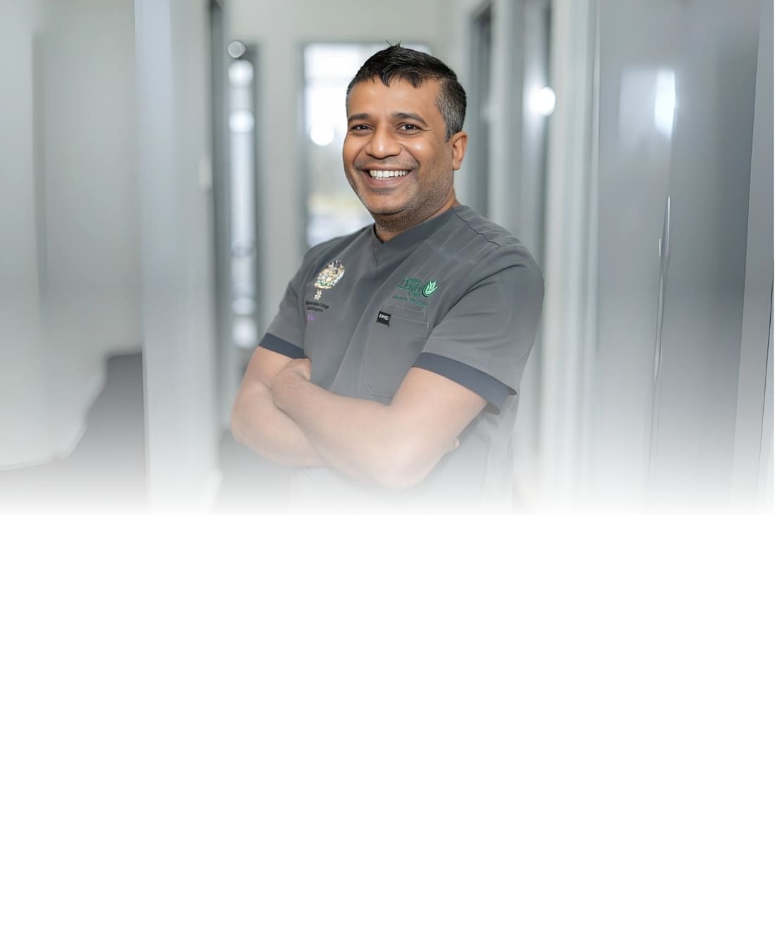 Professional male dentist in grey scrubs smiling confidently in the clinical hallway at Dandenong Dental Care.